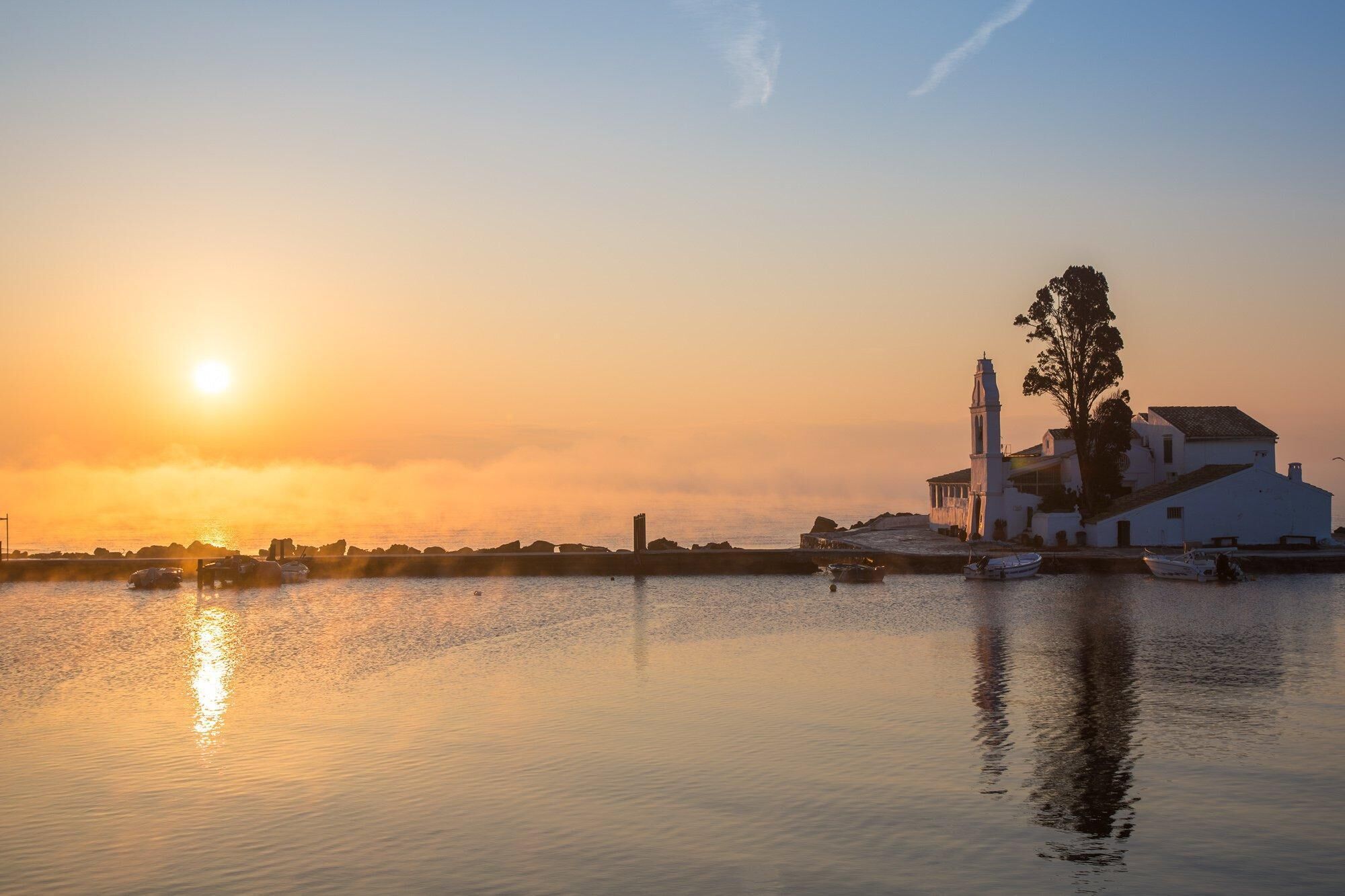 Eine Küstenlandschaft mit einem weißen Gebäude, einem Kirchturm und Booten auf ruhigem Wasser bei Sonnenuntergang.