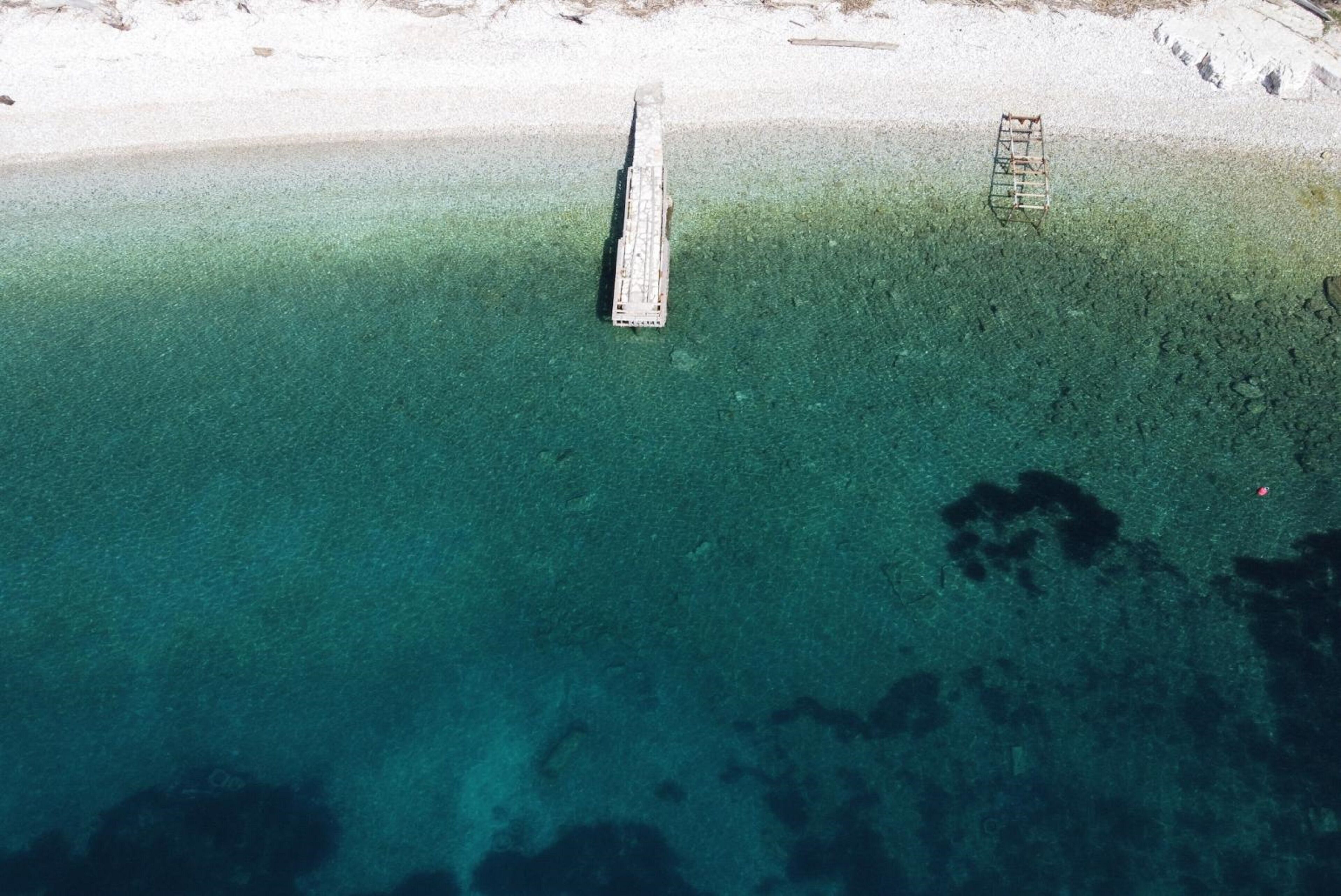 Een steiger die zich uitstrekt in een heldere blauwe zee, met een zandstrand en een ladder aan de rechterkant.