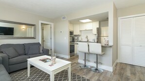 A compact living space with a grey sofa, a white coffee table, and a kitchen area featuring a microwave, oven, and refrigerator.