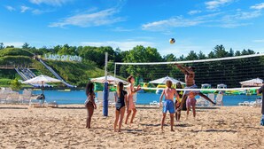 Een strandvolleybalwedstrijd op een zandstrand, met een waterpartij op de achtergrond, parasols en stoelen.