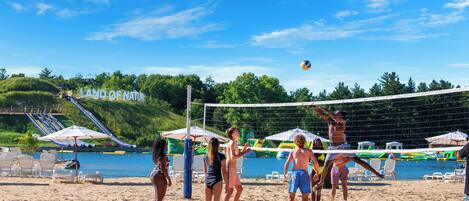 Partido de voleibol de playa en una arena, con un cuerpo de agua al fondo, sombrillas y sillas.
