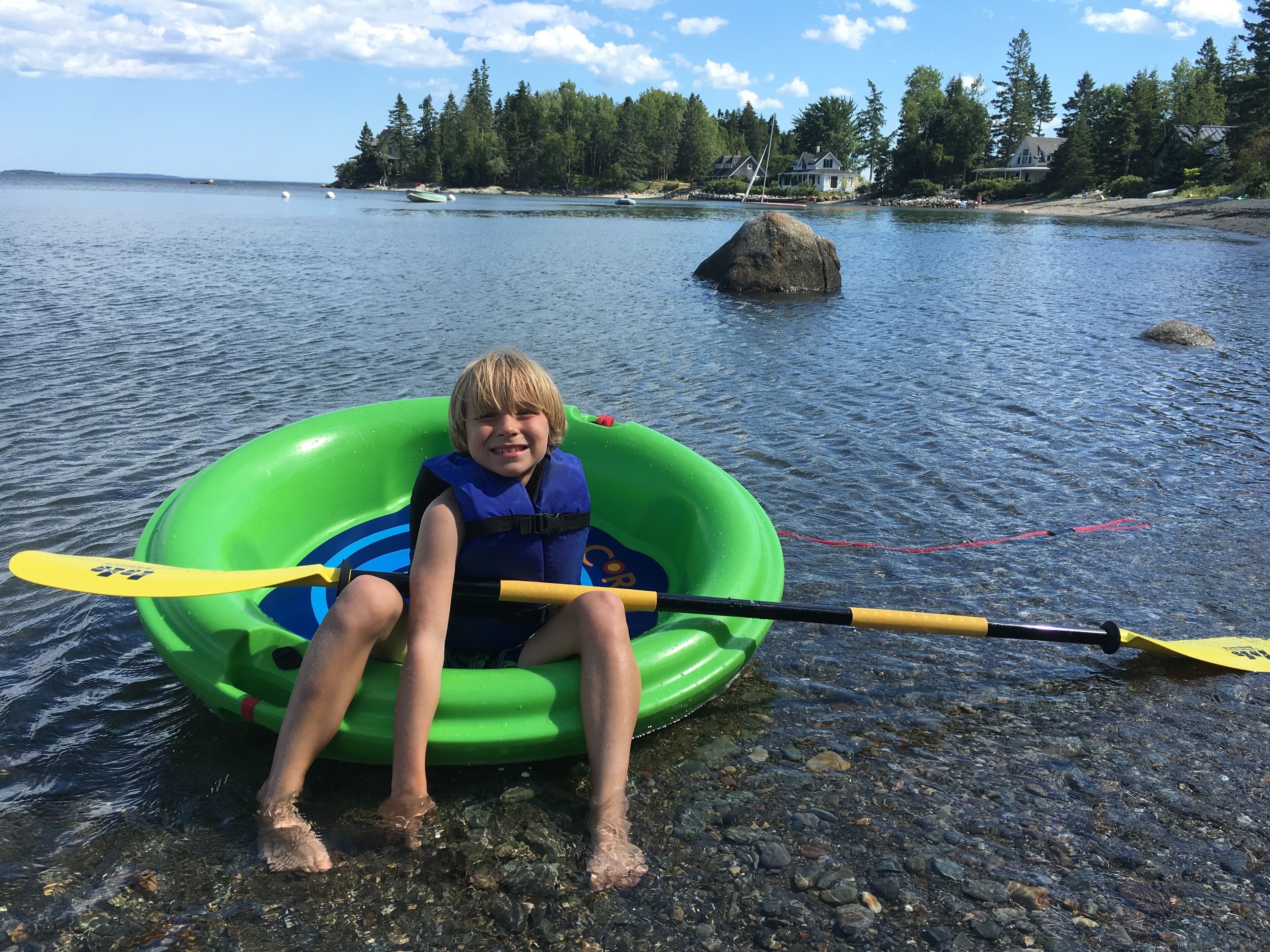 A person sitting in a green inflatable raft on a rocky shore with a body of water and houses in the background.