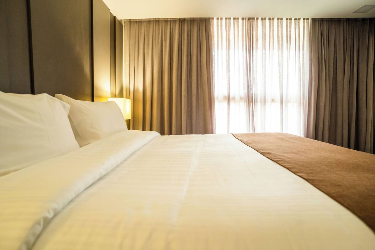Une chambre d’hôtel avec un grand lit, une lampe de chevet et une vue sur l’extérieur à travers des rideaux transparents.