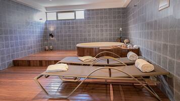A modern spa room with a wooden floor, a hot tub, and two massage tables with towels.