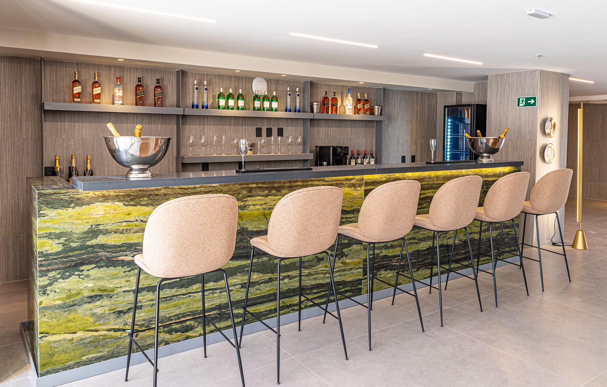 Zona de bar con mostrador de mármol, taburetes y una variedad de botellas de licor.