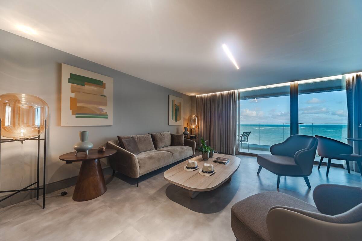 Un salon moderne avec un canapé, une table basse et des fauteuils, une grande fenêtre donnant sur la mer et des œuvres d’art abstraites accrochées au mur.