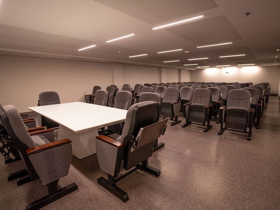 A conference room with a long table and rows of chairs, equipped with overhead lighting.