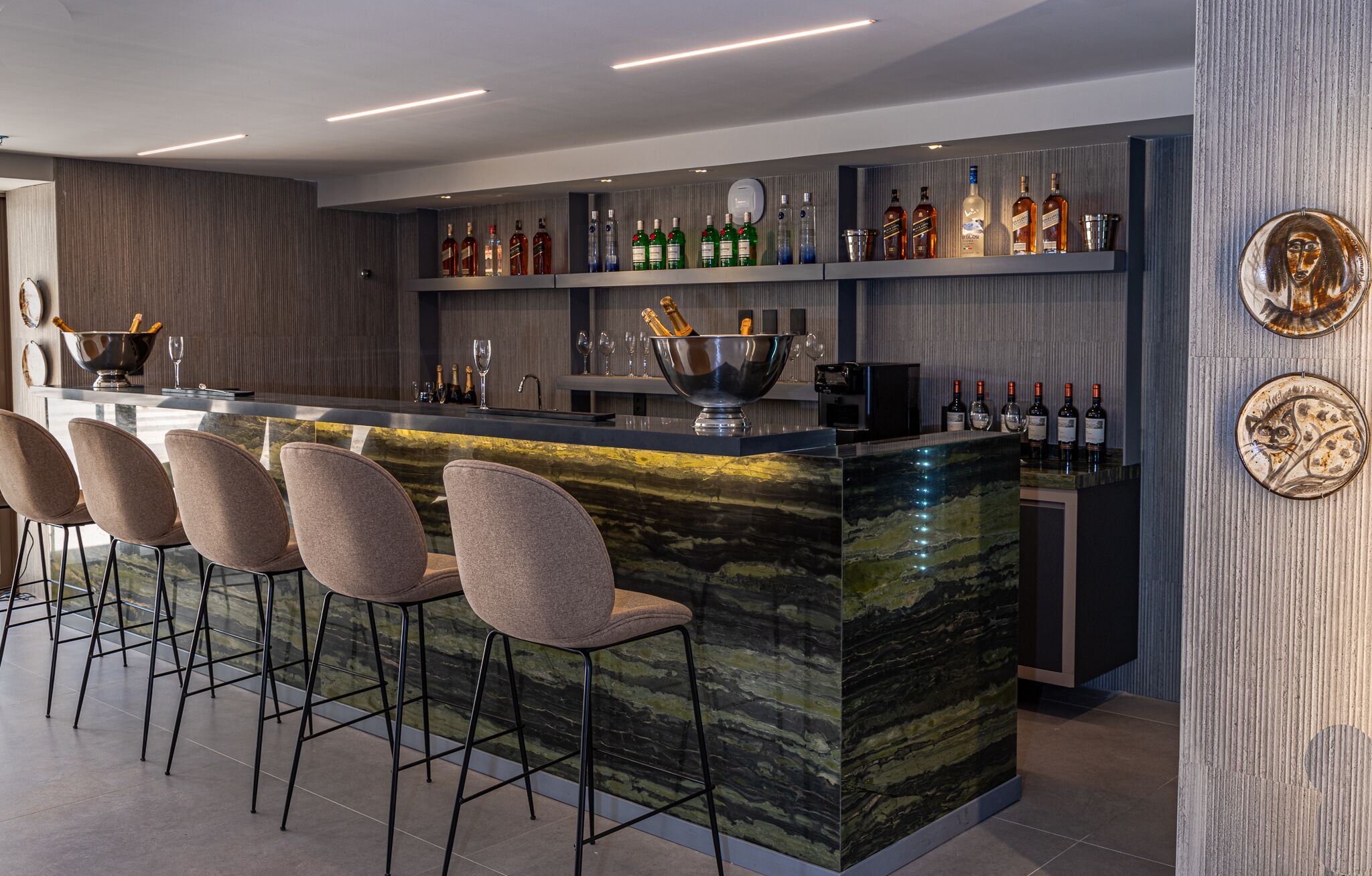 A modern bar area with a marble countertop, bar stools, and a variety of liquor bottles.