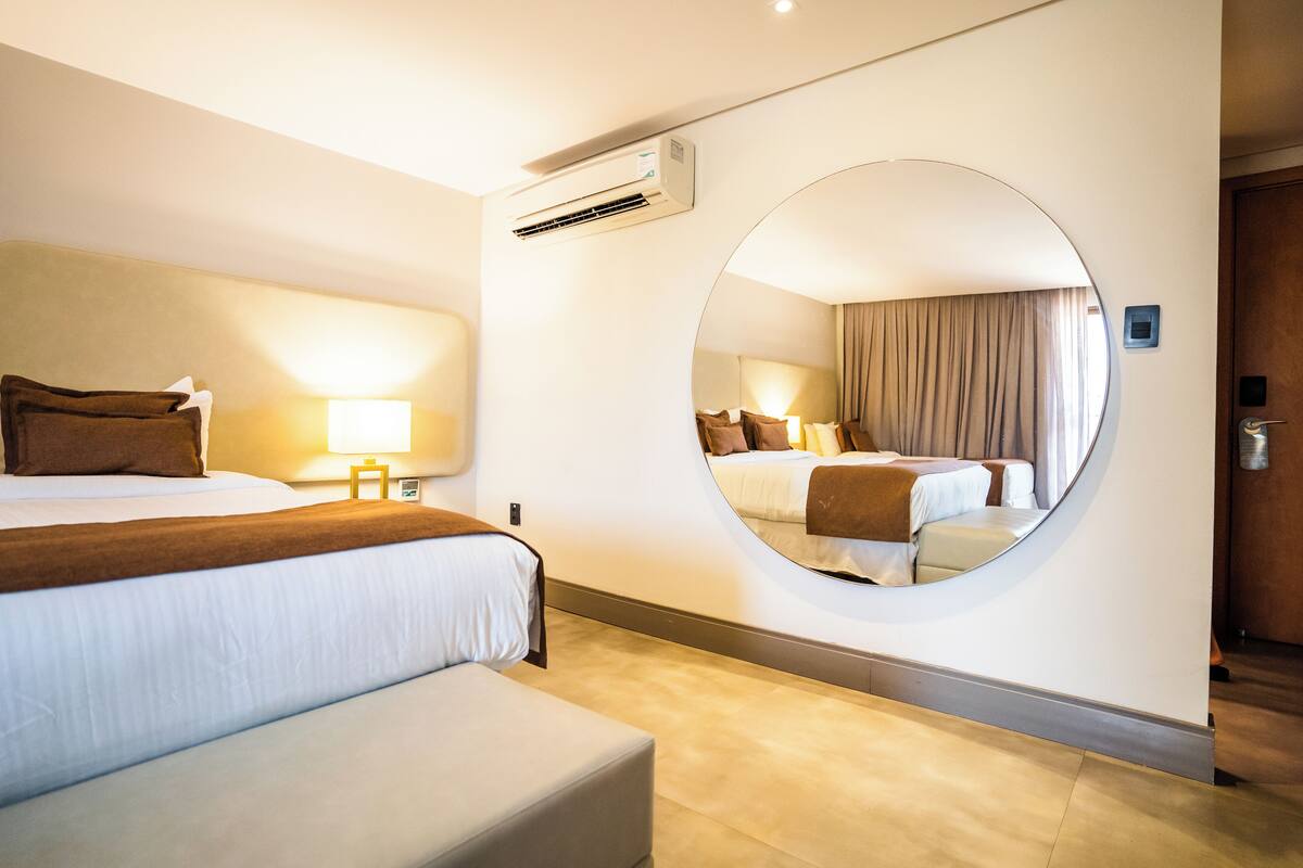 Une chambre d’hôtel avec un grand lit, une table de chevet avec une lampe, un miroir rond au mur et un appareil de climatisation.