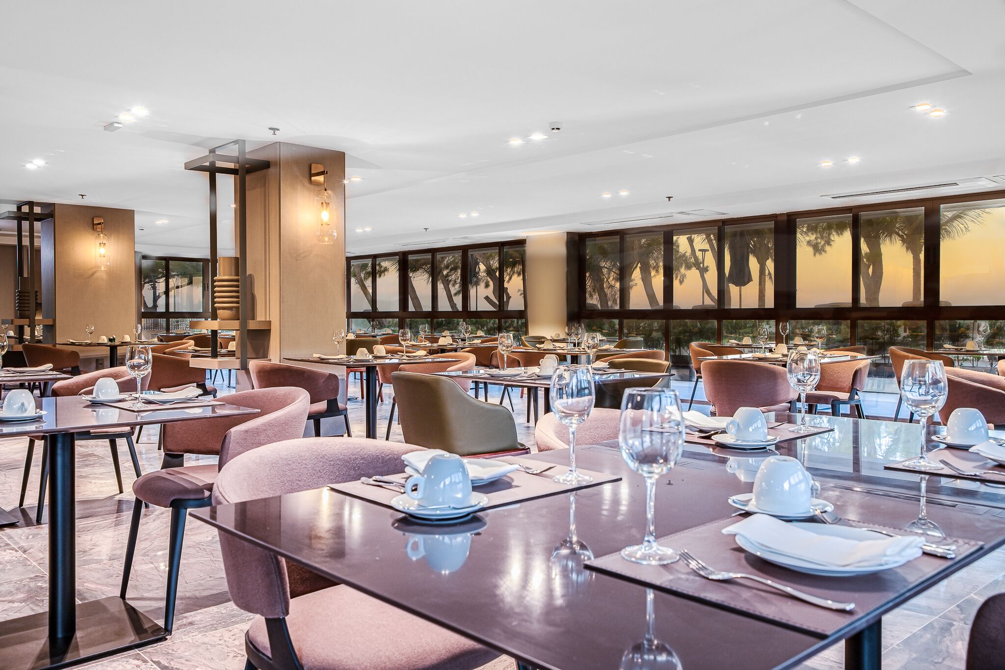 Un vaste espace dédié à la salle à manger, avec de nombreuses tables dressées pour le service, de grandes fenêtres offrant une vue sur les palmiers et un design de plafond moderne.