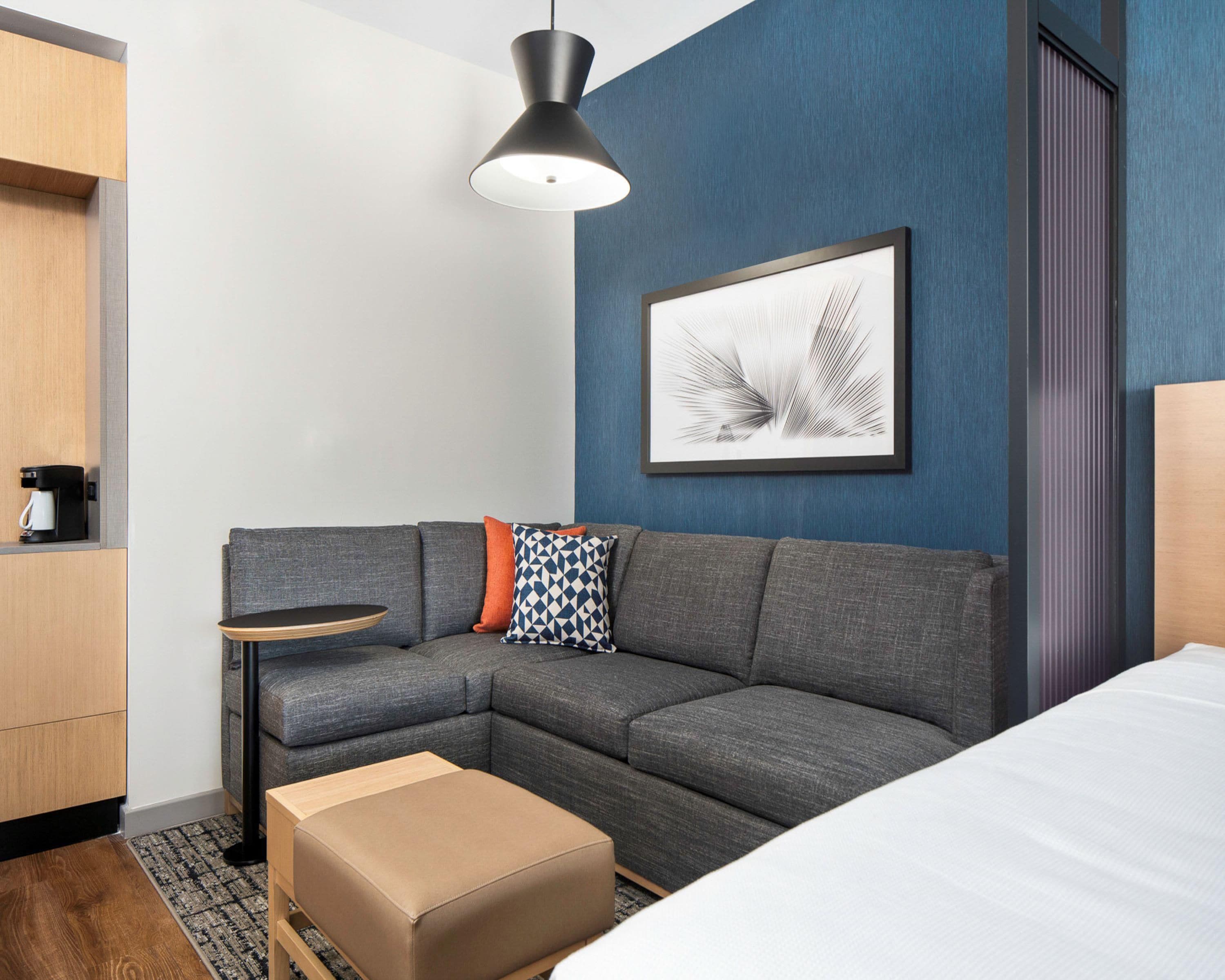 A modern hotel room with a gray sectional sofa, a small side table, a coffee machine, a framed abstract art piece, and a wooden wardrobe.
