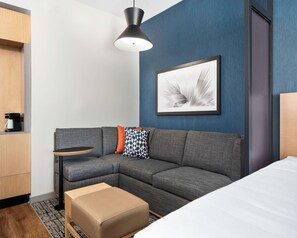 A modern hotel room with a gray sectional sofa, a small side table, a coffee machine, a framed abstract art piece, and a wooden wardrobe. - Hyatt Place Allentown / Lehigh Valley (Allentown)