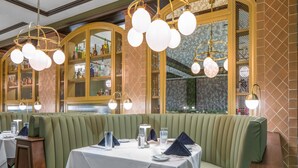 Een restaurant met groen bekleden zitbanken, witte tafelkleden en hangende, bolvormige lampen.