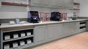 A commercial kitchen with two coffee machines, a display case with pastries, and neatly arranged white plates and bowls.