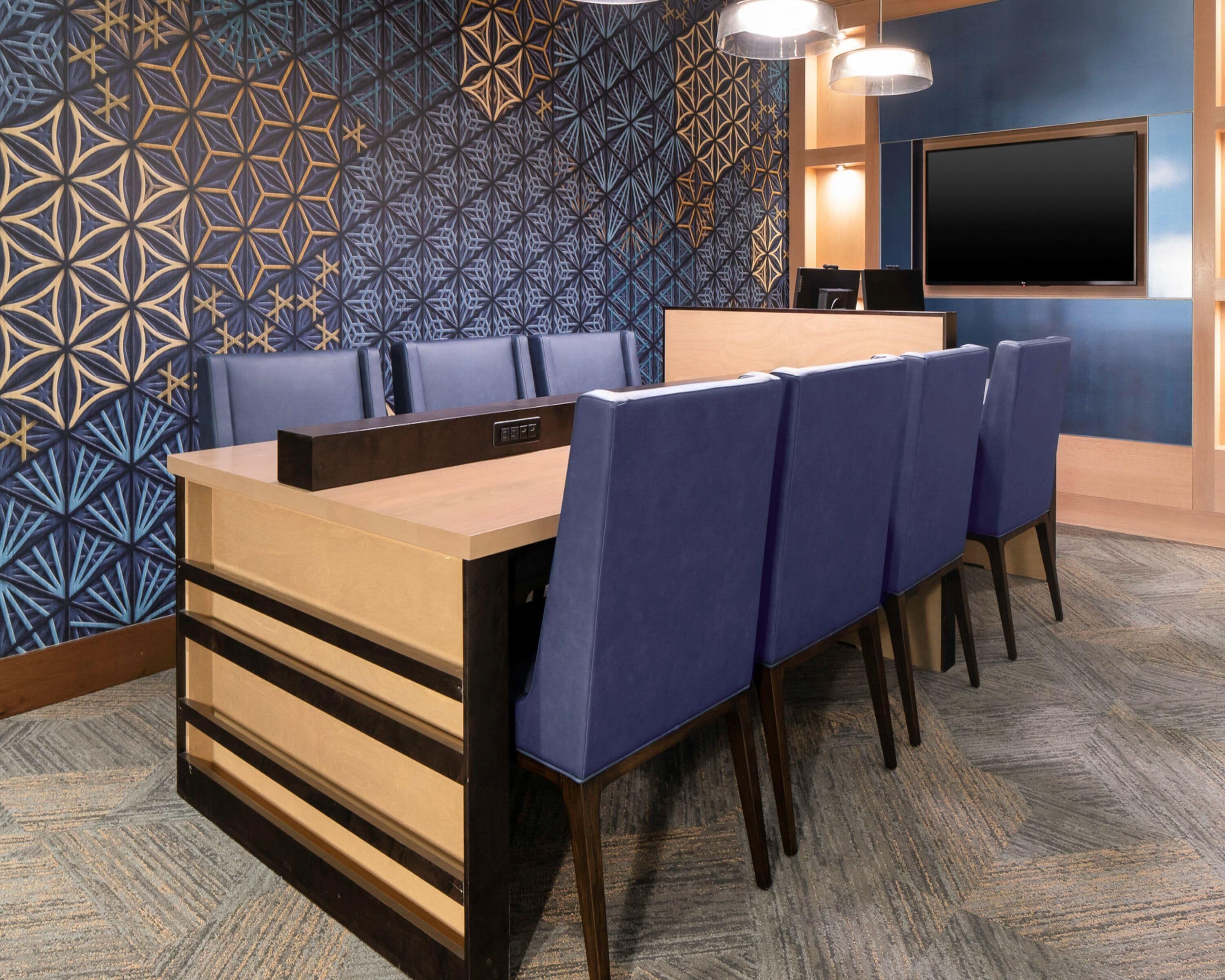 Un salón de conferencias con una mesa larga, sillas azules, un escritorio de madera, un televisor de pantalla plana y decoración de pared estampada.