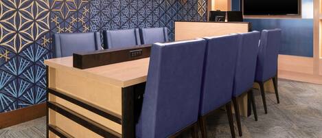 Un salón de conferencias con una mesa larga, sillas azules, un escritorio de madera, un televisor de pantalla plana y decoración de pared estampada.