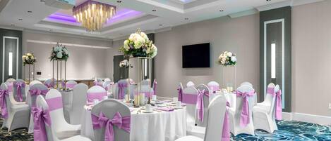 Un salón de banquetes con mesas redondas, sillas blancas con cintas rosas y centros de mesa florales.
