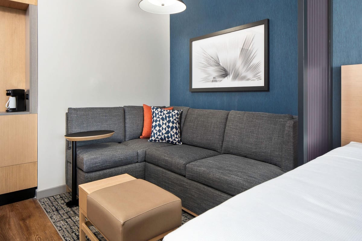 Una habitación de hotel moderna con un sofá seccional gris, una mesita redonda pequeña, una cafetera, una obra de arte enmarcada y una cama grande.