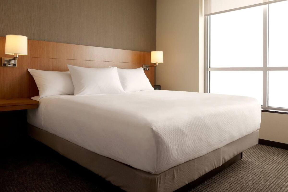 Habitación de hotel con una cama grande, dos lámparas de noche, un cabecero de madera y un ventanal amplio.