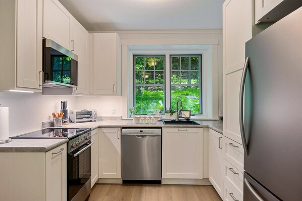 A modern kitchen with white cabinets, stainless steel appliances, a dishwasher, and a window with a view of greenery.
