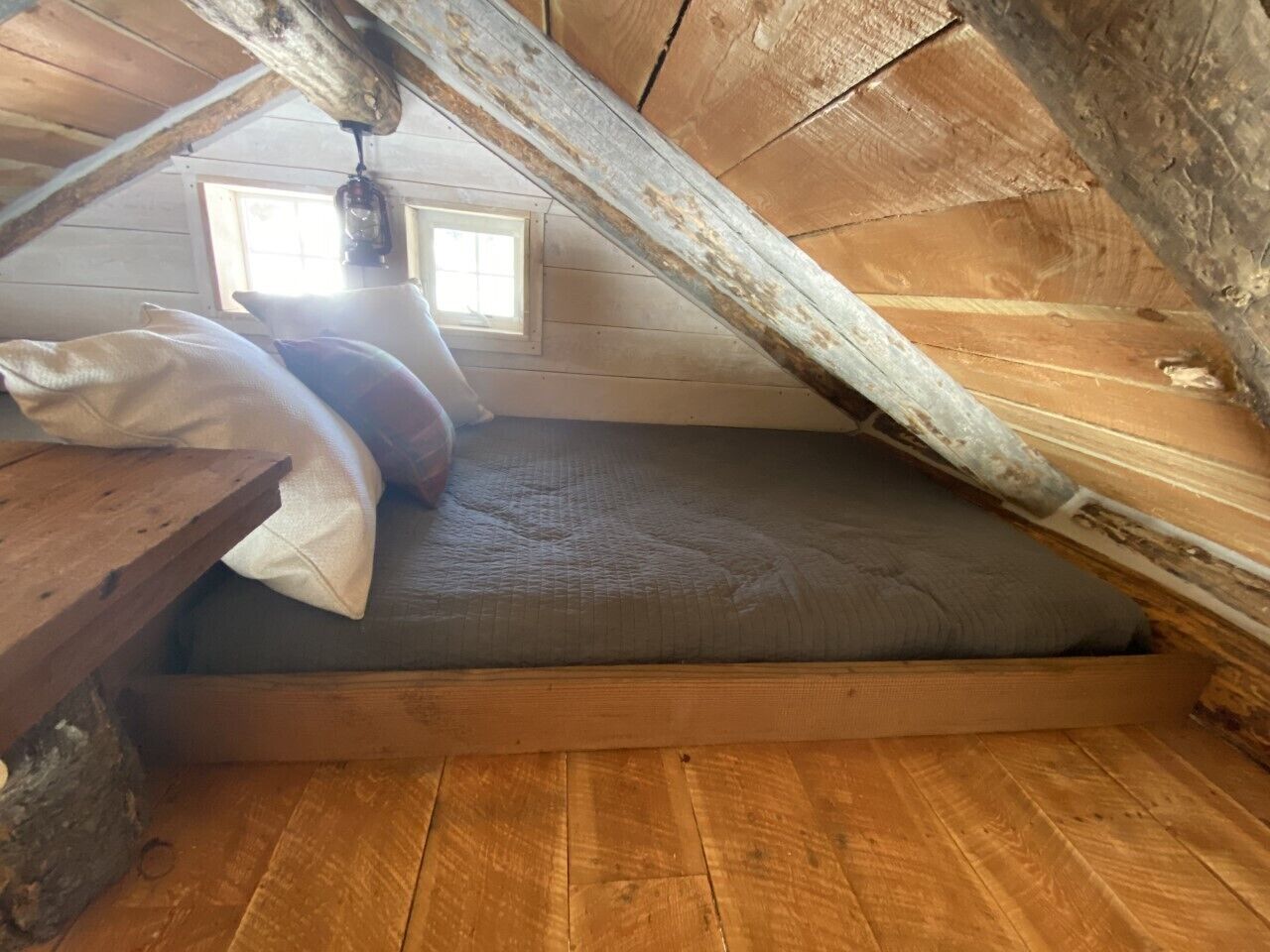 Et soveværelse med skråvægge og trægulv, en madras og en hængende lampe.
