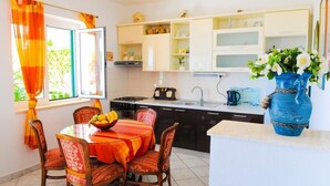 Una cocina con armarios blancos, una mesa de comedor con sillas, un jarrón azul con flores y una ventana con cortinas naranjas.