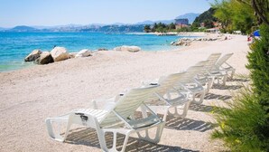 Una playa con sillas de descanso blancas alineadas sobre la arena, un mar azul cristalino y un paisaje urbano distante.