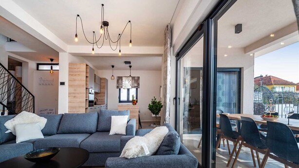 A modern living room with a grey sofa, a small table, and a dining area with chairs and a glass door leading outside.