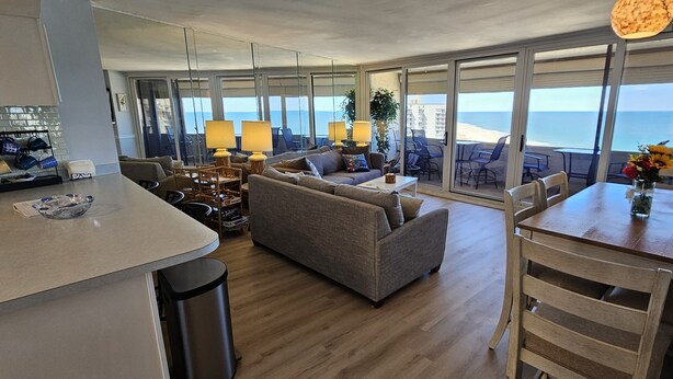 Ampla sala de estar com sofá, mesa de jantar e cadeiras, área de cozinha com balcão e banquetas, e grandes portas de vidro deslizantes que dão para uma varanda com vista para o mar.