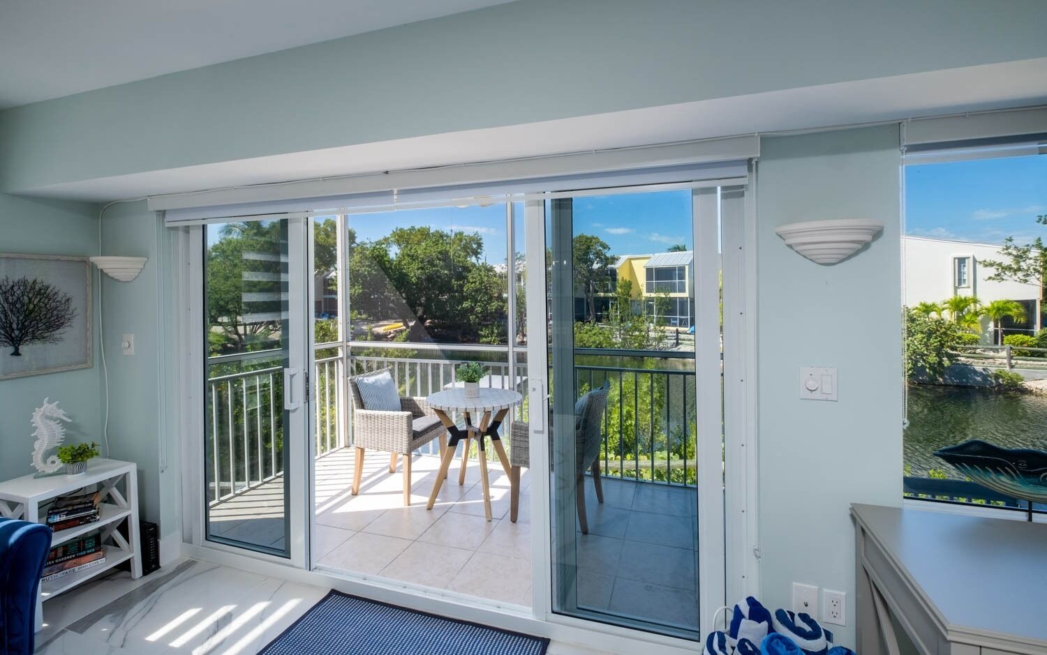 A modern apartment with a sliding glass door leading to a balcony with a table and chairs, a small bookshelf, and a decorative plant.