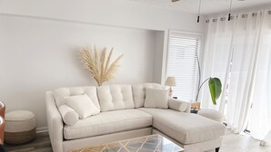 Una sala de estar con un sofá seccional color beige, una mesa de centro de vidrio y un ventanal con cortinas blancas.