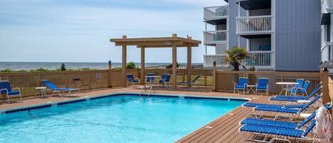 A swimming pool with blue lounge chairs and a wooden deck, surrounded by a wooden fence and a building with balconies.