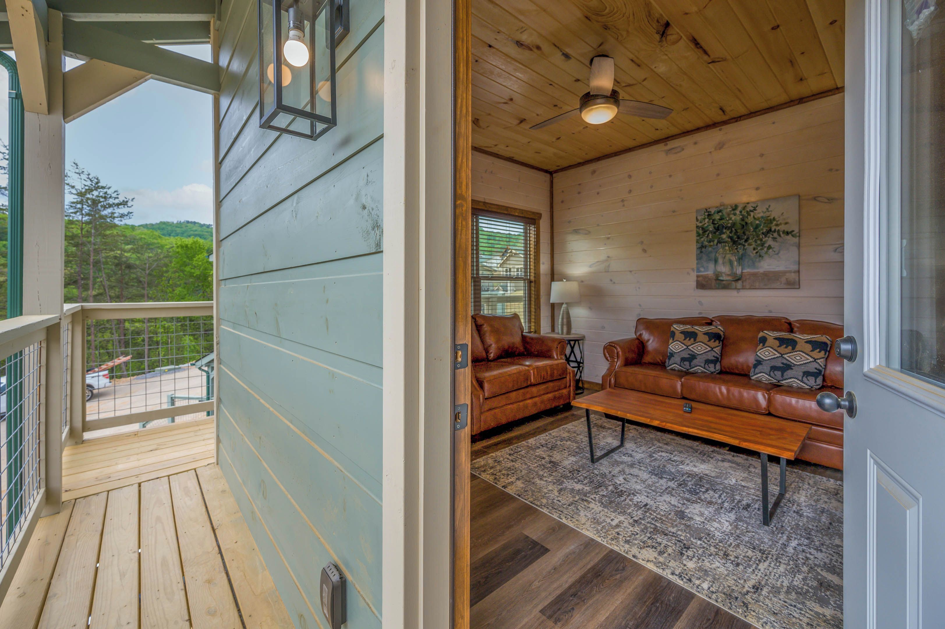 A wooden deck with a view of trees, a brown leather sofa with patterned cushions, and a ceiling fan in a room with wooden walls and a window.