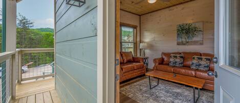 A wooden deck with a view of trees, a brown leather sofa with patterned cushions, and a ceiling fan in a room with wooden walls and a window.