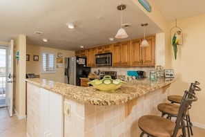 A kitchen with wooden cabinets, a granite countertop, a microwave, and a green decorative bowl. There are three bar stools and a hanging bird ornament. - Scenic 2 Bed Condo by the Beach with Pool - Perfect View (St. Augustine)