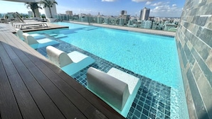 Una piscina infinita en la azotea con agua cristalina y azul, rodeada de una terraza de madera y sillones de descanso, con vistas a un paisaje urbano con edificios modernos.