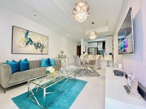A modern living room with a grey sofa, blue cushions, a glass coffee table, and a chandelier.