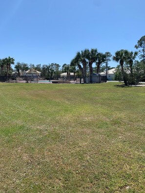 A grassy area with a fenced pool, palm trees, and a building with a thatched roof.