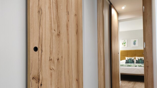 Un couloir mène à une chambre dotée d’une porte en bois, d’un lit avec une tête de lit jaune et d’un mur orn é de deux tableaux encadrés.