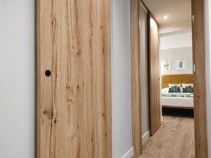 Ein Flur, der zu einem Schlafzimmer mit Holztür, einem Bett mit gelbem Kopfteil und einer Wand mit zwei gerahmten Bildern führt.