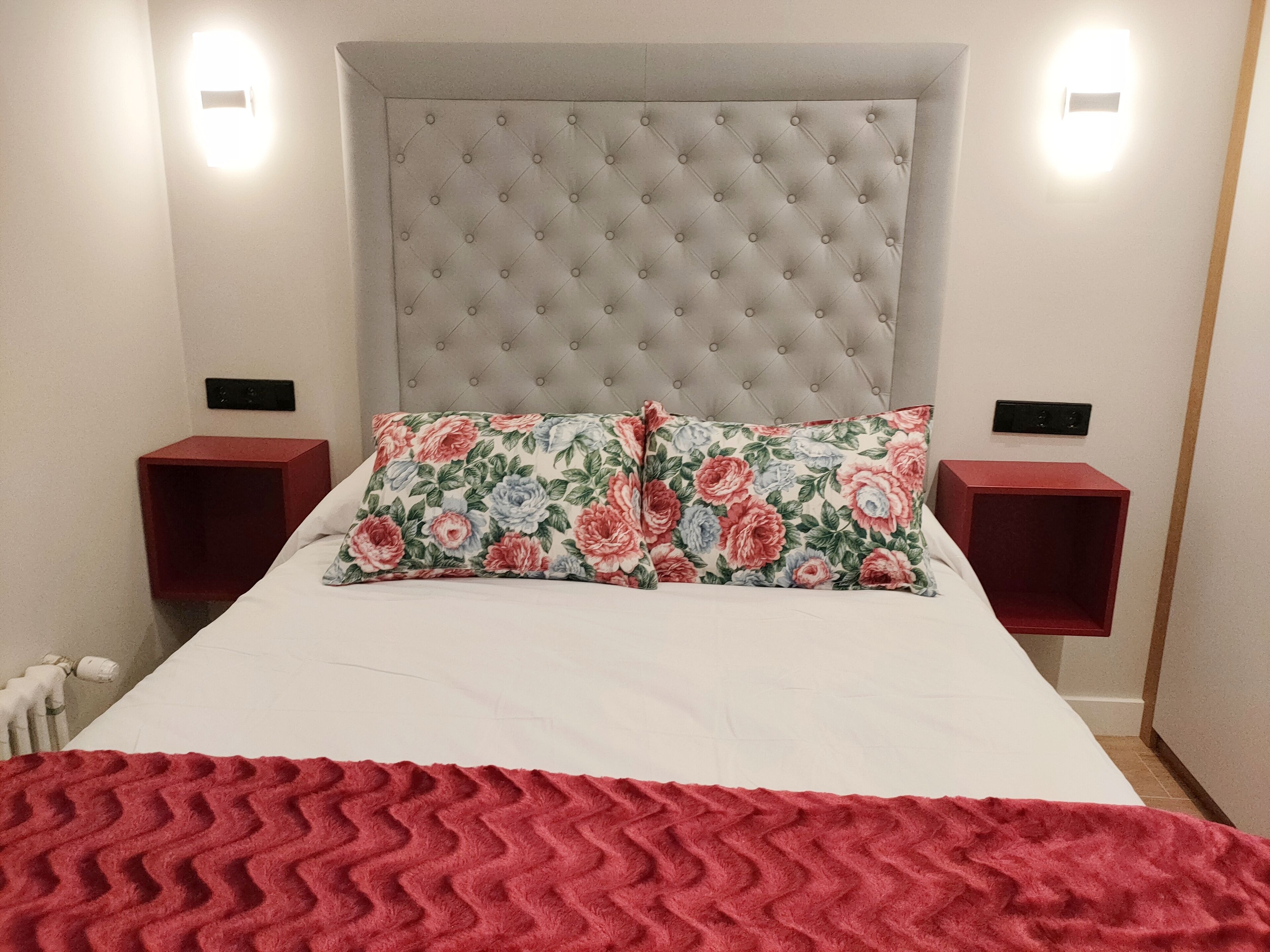 Una cama bien tendida con un edredón rojo, un cojín estampado y un cabecero capitonado. Hay dos mesitas de noche con bases rojas y interruptores negros.