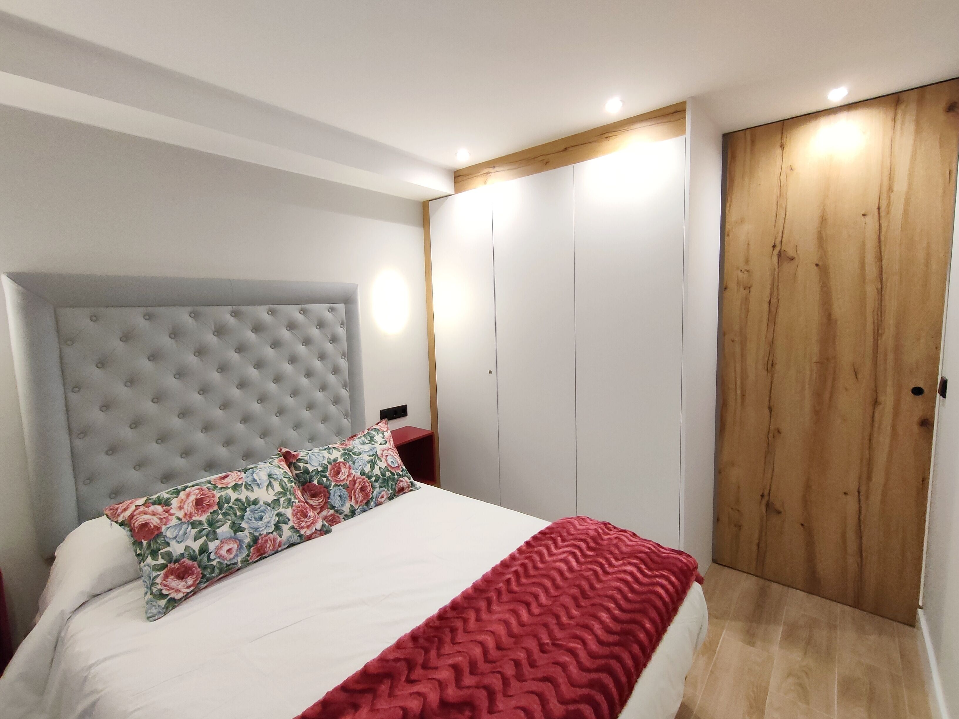 Une chambre à coucher avec une tête de lit capitonnée, un coussin à motifs floraux, une couverture rouge, une armoire en bois et une table de chevet avec une lampe.