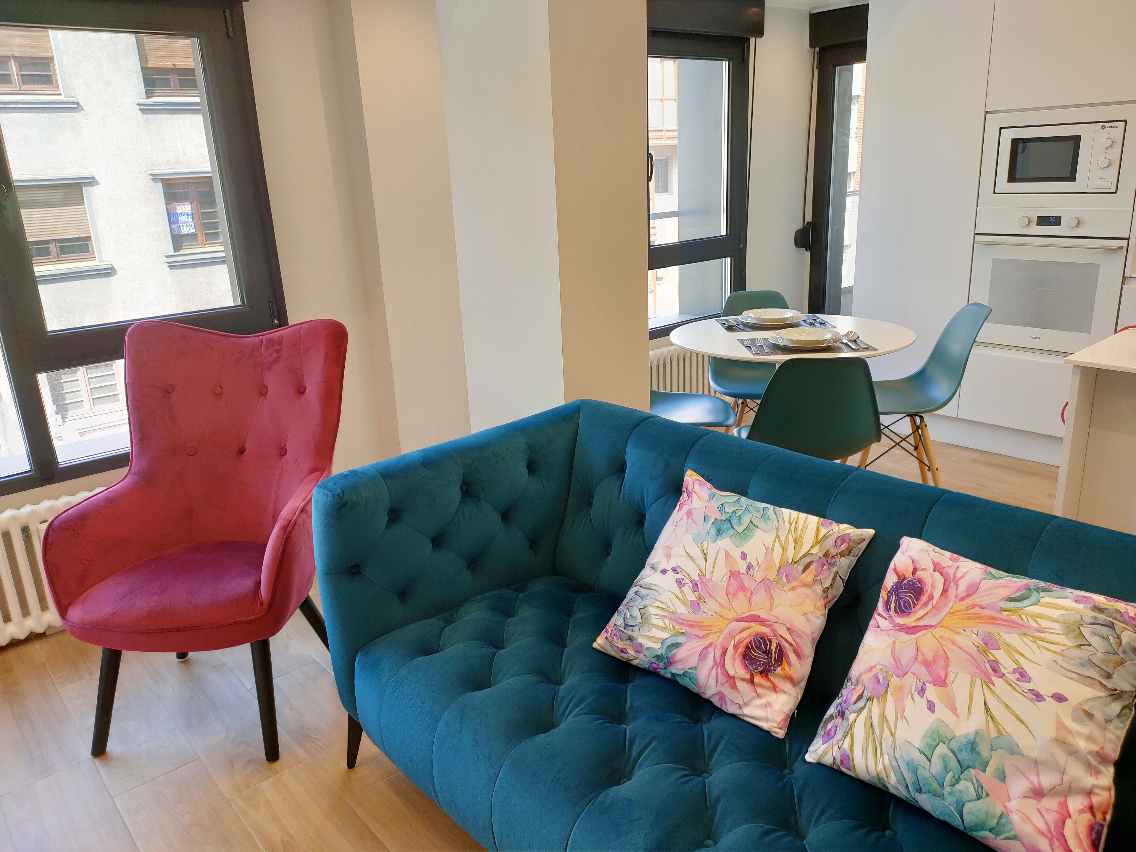 A modern living room with a teal sofa, a red armchair, and a dining area with a table and chairs.