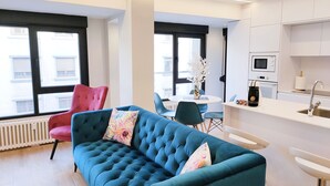 Un salon moderne avec un canapé bleu et un fauteuil rose, un coin repas avec une table et des chaises, et une cuisine avec des meubles blancs et des appareils électroménagers.