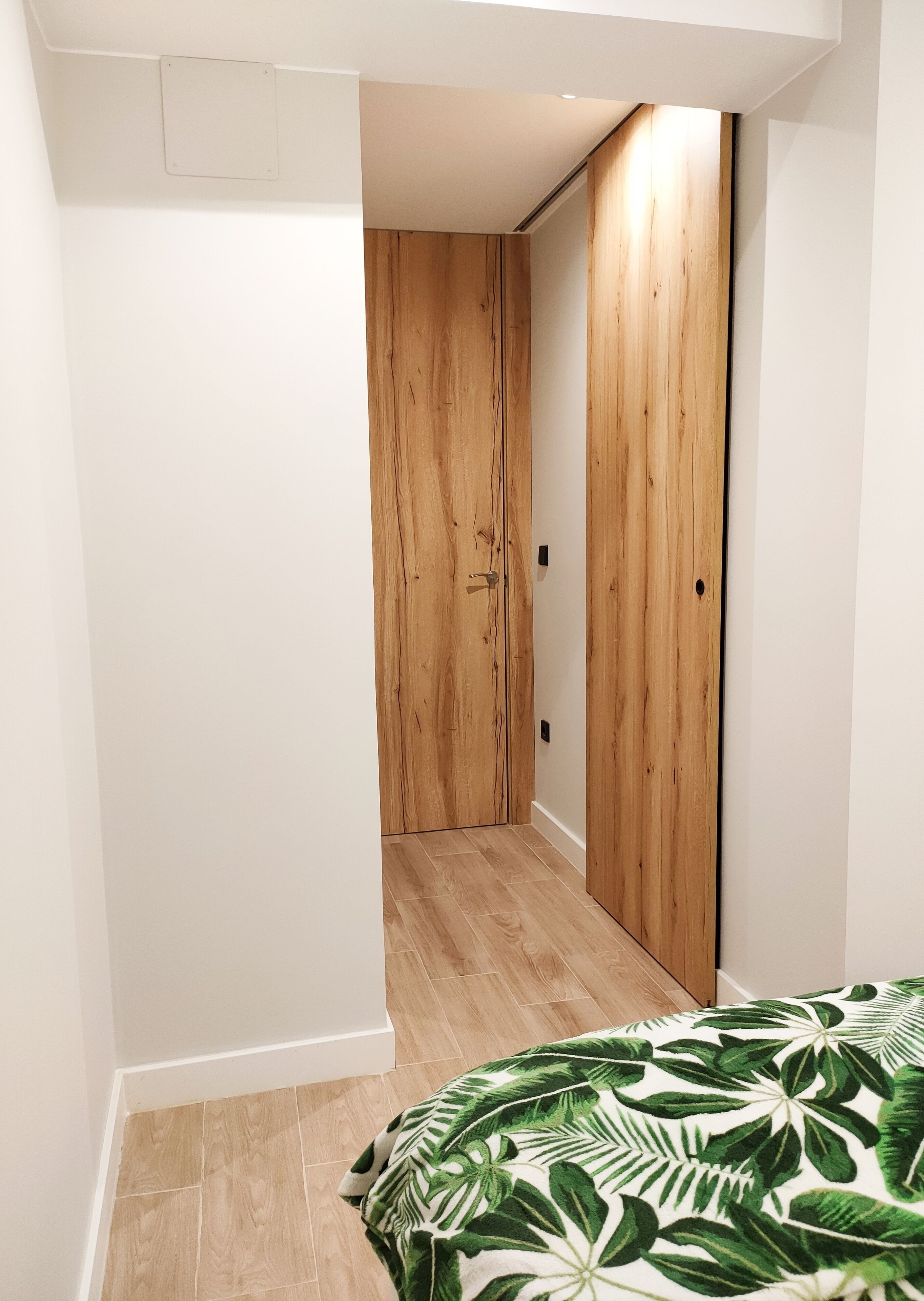 Um corredor estreito com portas de madeira e uma cama com colcha estampada em folhas verdes.