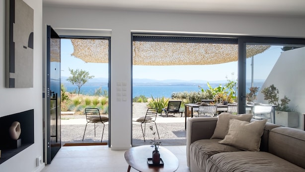 Un salon moderne avec un canapé, une table basse et une vue sur une plage et des montagnes, grâce à de grandes fenêtres.