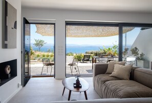 Un salon moderne avec un canapé, une table basse et une vue sur une plage et des montagnes, grâce à de grandes fenêtres.