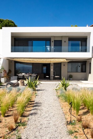Une maison moderne à deux étages, dotée d’une allée en gravier, d’un espace extérieur aménagé pour se détendre et d’une grande baie vitrée.