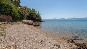 Une plage de galets bordée d’une eau cristalline, une falaise rocheuse et, au loin, une chaîne de montagnes.