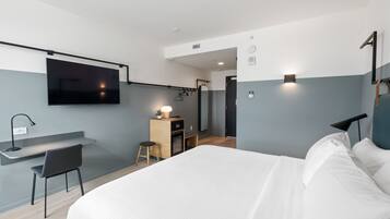 A modern hotel room with a bed, a desk, a TV, and a small kitchenette.
